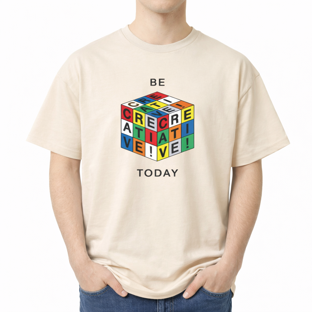 Beige t-shirt with colorful Rubik's Cube design and text on a white background. A flat lay of a short-sleeve t-shirt. The center chest features a graphic of a 3D puzzle cube with brightly colored grid squares in red, blue, green, yellow, orange, and white. The black letters across the visible grids spell out "CREATIVE!". Above the cube is the text "BE" and below the cube is the text "TODAY" in a simple, modern black font.
