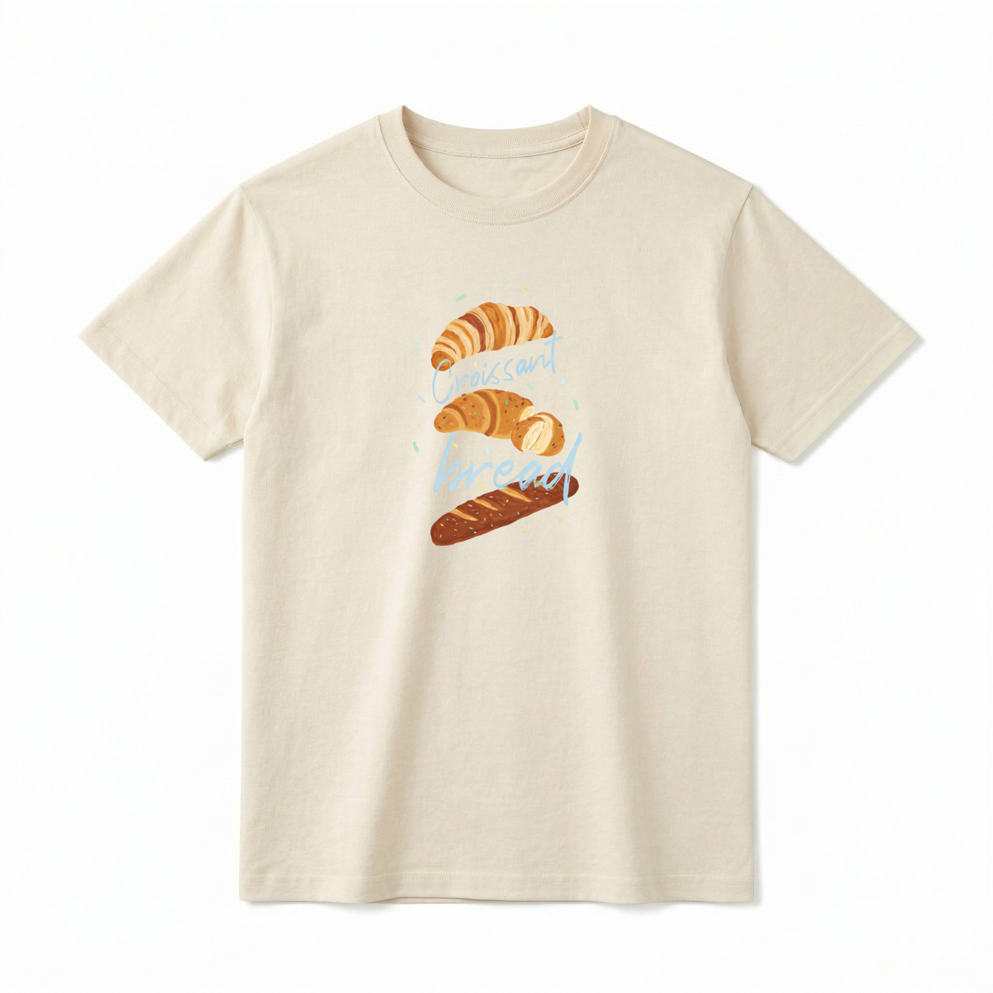 A flat lay of a short-sleeve t-shirt. The center chest features a hand-drawn graphic of vertically stacked baked goods: a golden croissant at the top, a sliced pastry in the middle, and a seeded baguette at the bottom. Light blue script text reading "Croissant" and "bread" is woven playfully across and between the pastries, surrounded by tiny, colorful confetti accents.