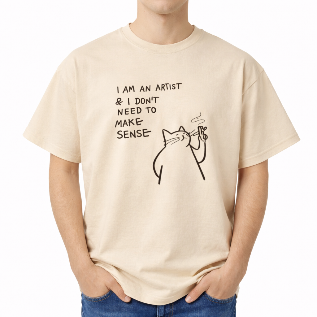 Beige t-shirt with a cat graphic and text worn by a person on a white background. The center chest features a black minimalist line-art graphic of a smug, chunky cat holding a smoking object. To the left of the cat, playful handwritten-style text reads "I AM AN ARTIST & I DON'T NEED TO MAKE SENSE" stacked in four lines.